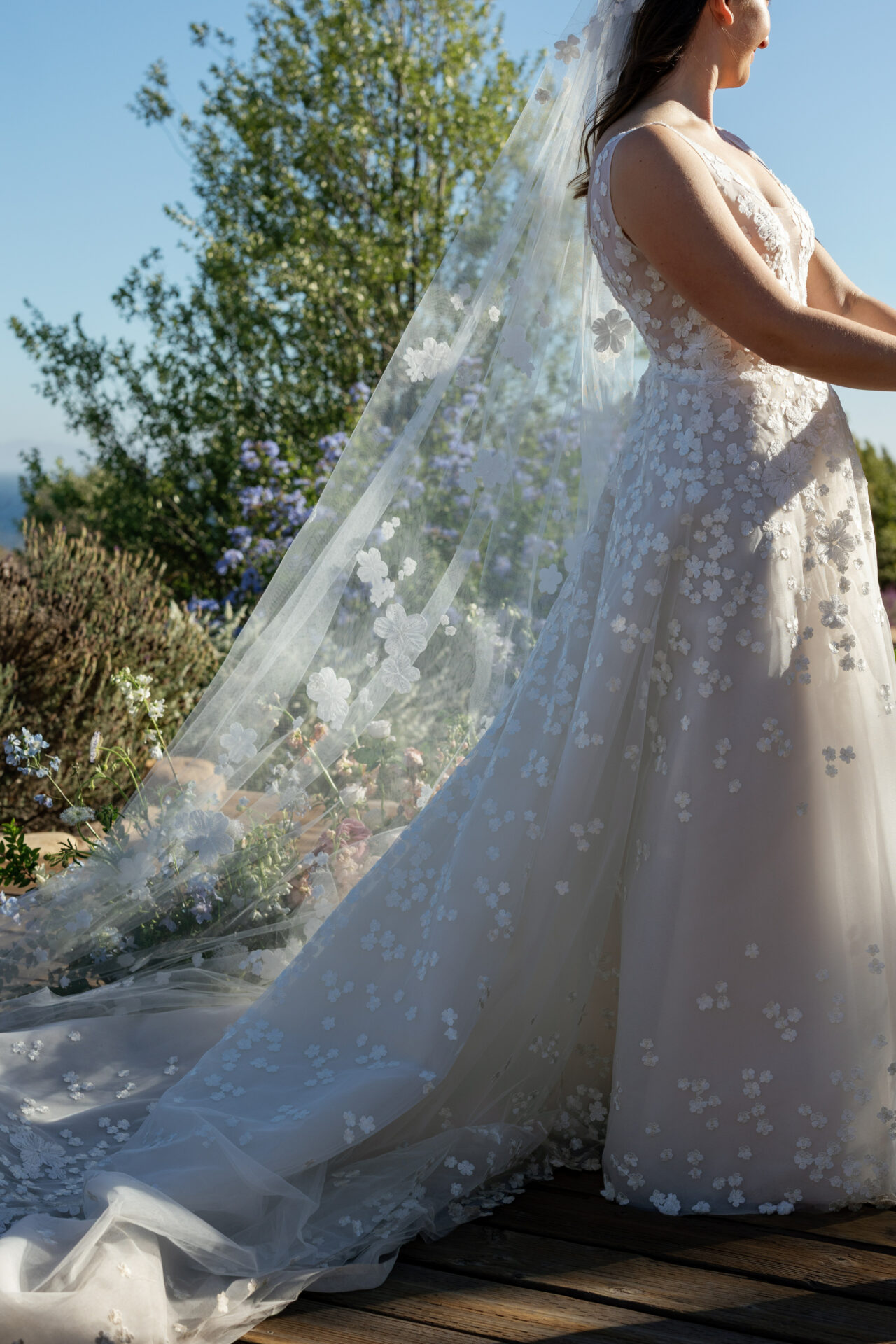 Bride in floral gown on deck, sunlight, greenery.