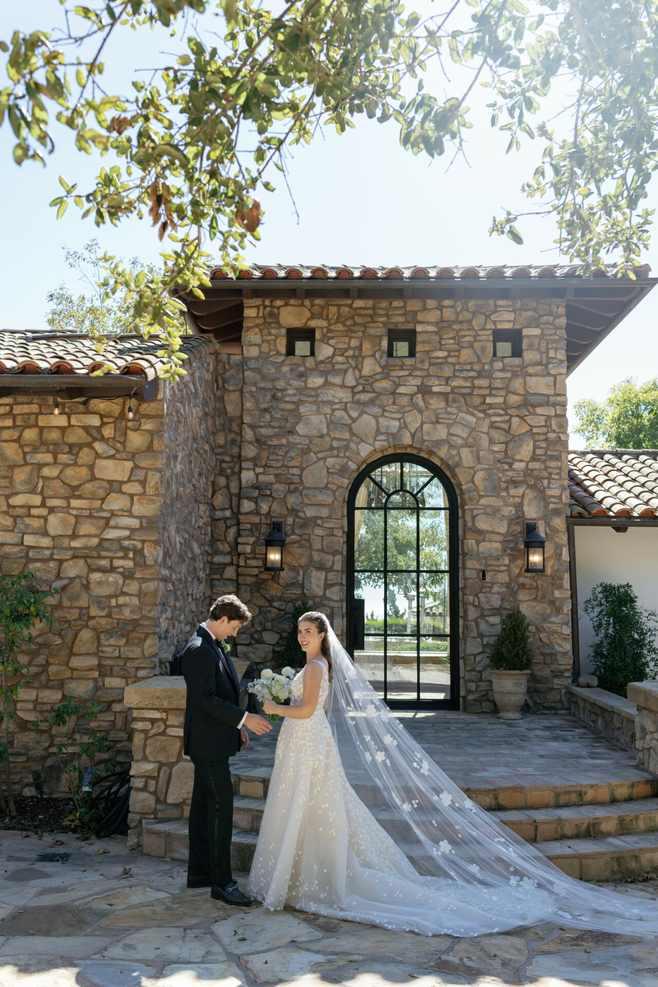 Bride and groom smile, holding hands outside sunlit stone building.