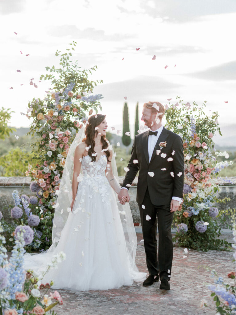 Bride and groom hold hands under floral arch at sunset.