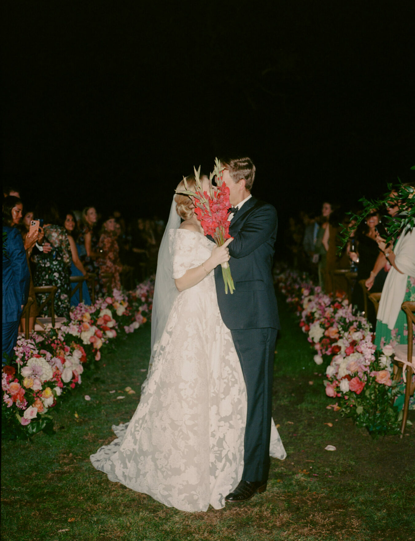 Bride and groom kiss outdoors at night among florals.
