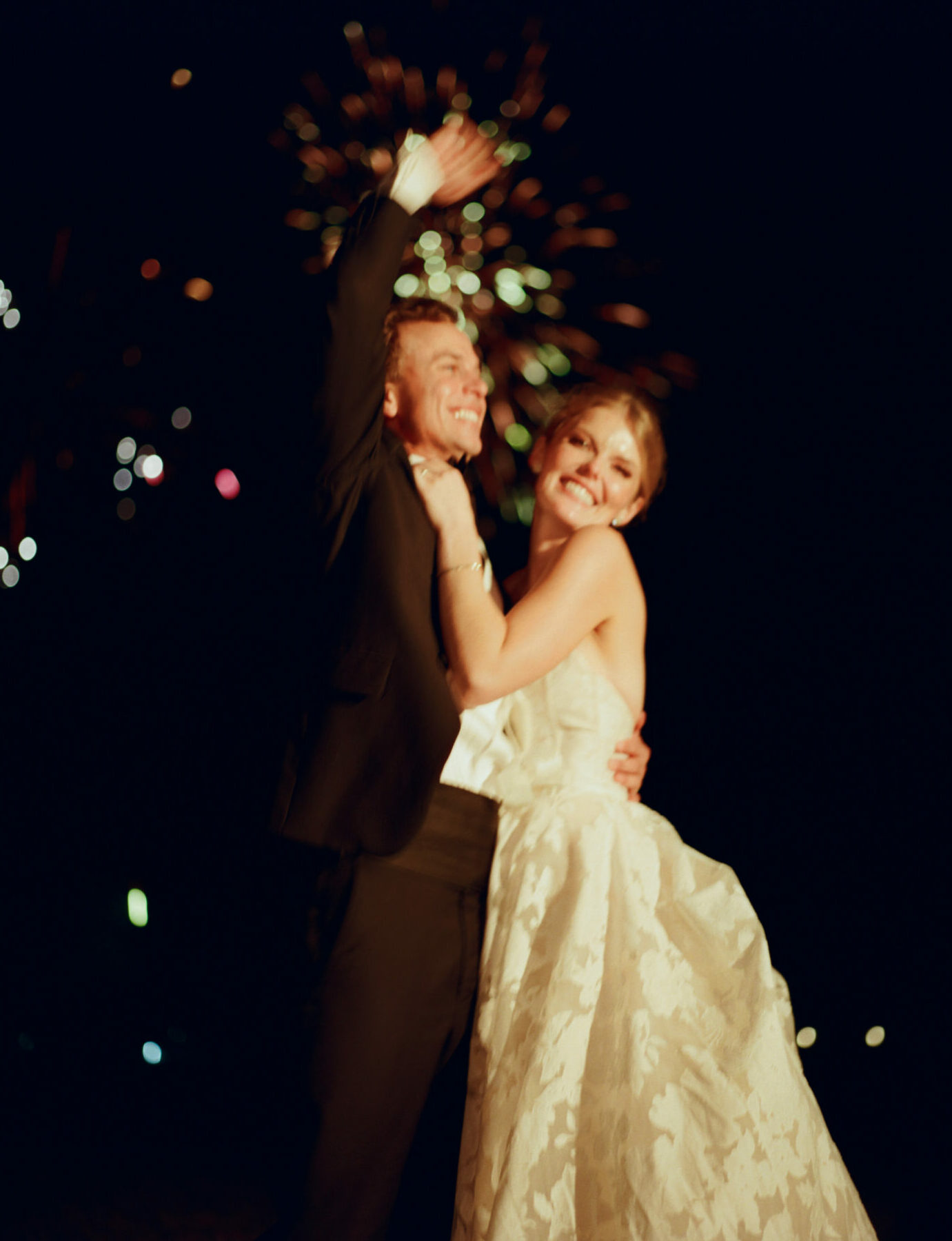 Couple in formalwear embrace under fireworks at night.