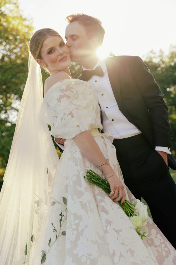 Bride in Mira Zwillinger gown kissed on cheek by groom, sunlight behind.