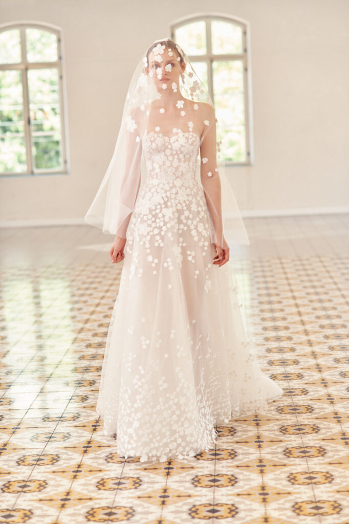 Bride in sheer floral gown stands on patterned floor.