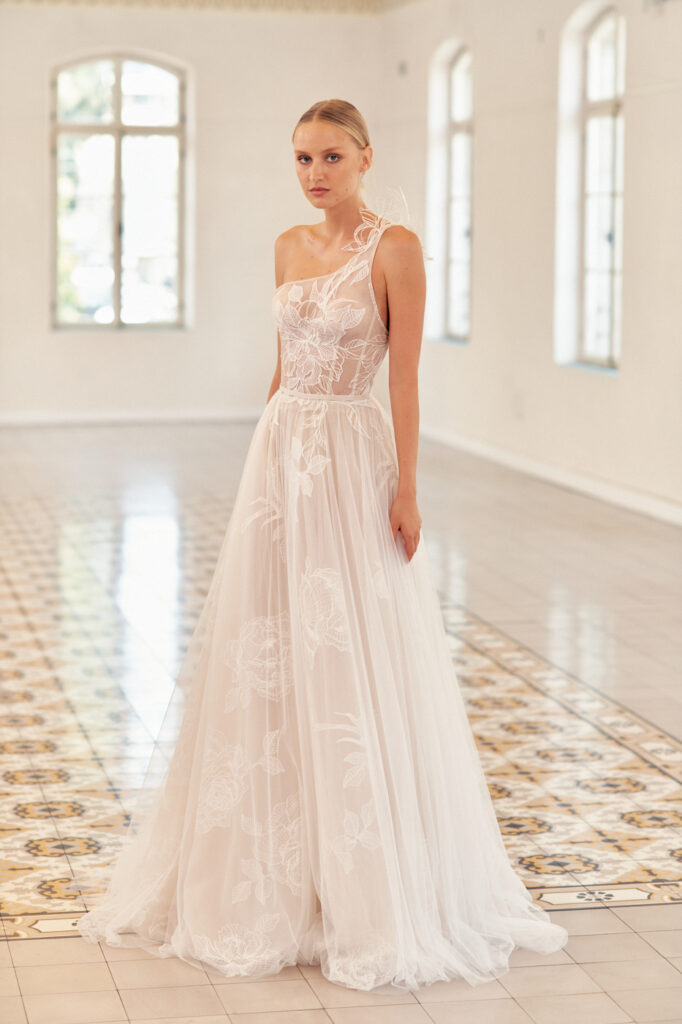 Bride in sheer one-shoulder floral dress, bright room.