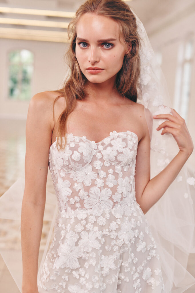 Woman in strapless floral lace wedding dress holds veil indoors.