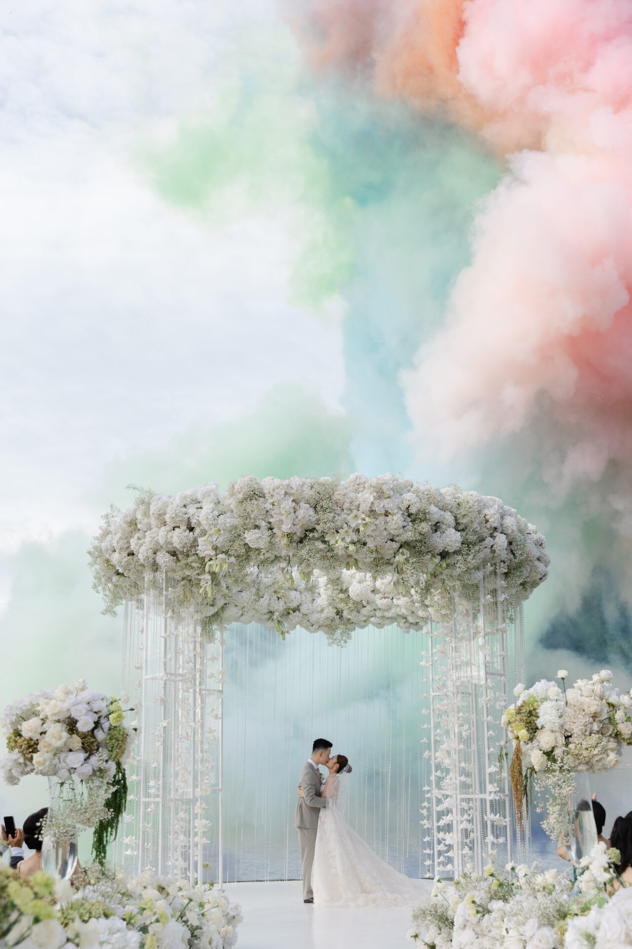 Bride and groom kiss beneath arch, smoke clouds behind.
