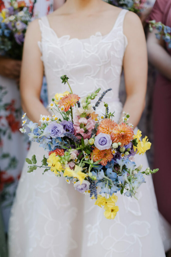 Bride in white floral dress holds colorful bouquet.