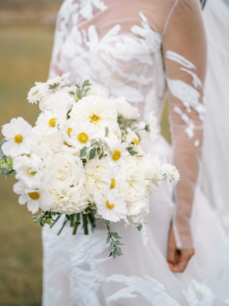 Bride in white gown holds bouquet on grassy field.