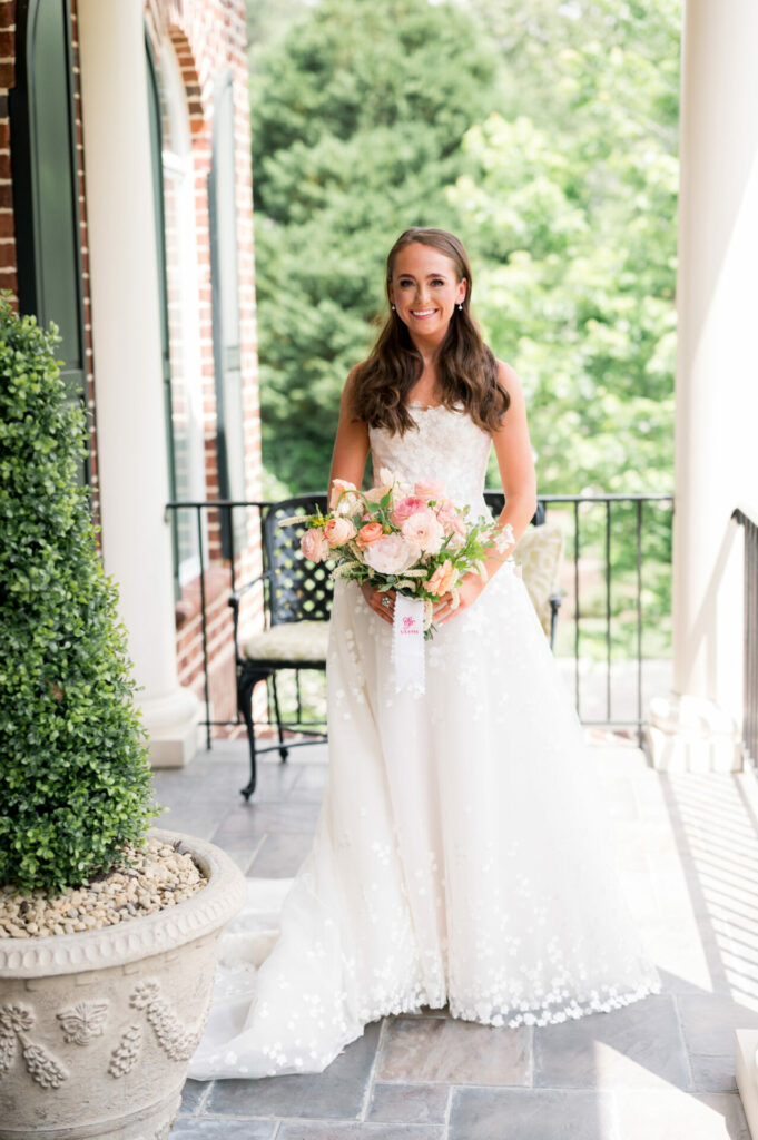 Katya Ruiz smiles on porch in wedding dress, bouquet.