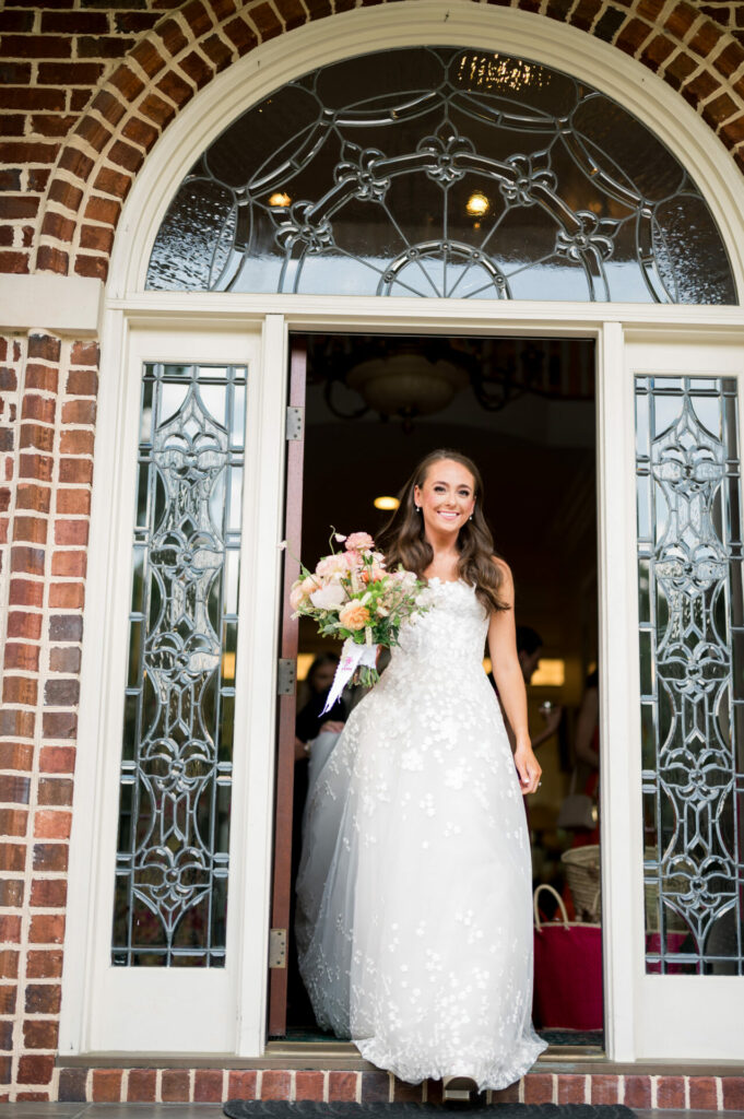 Katya Ruiz smiles in couture gown with bouquet by glass-paneled brick doorway.