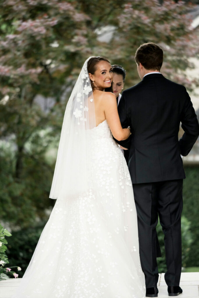 Bride in luxury gown with groom, pink blossoms blurred.
