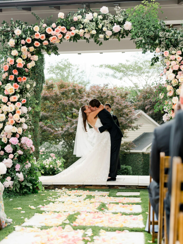 Bride and groom kiss beneath floral arch at wedding.
