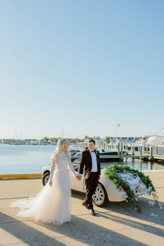 Bride and groom walk by vintage car, marina background.