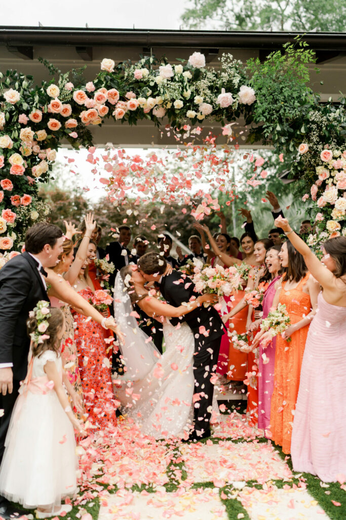 Couple kisses under floral arch as guests cheer.
