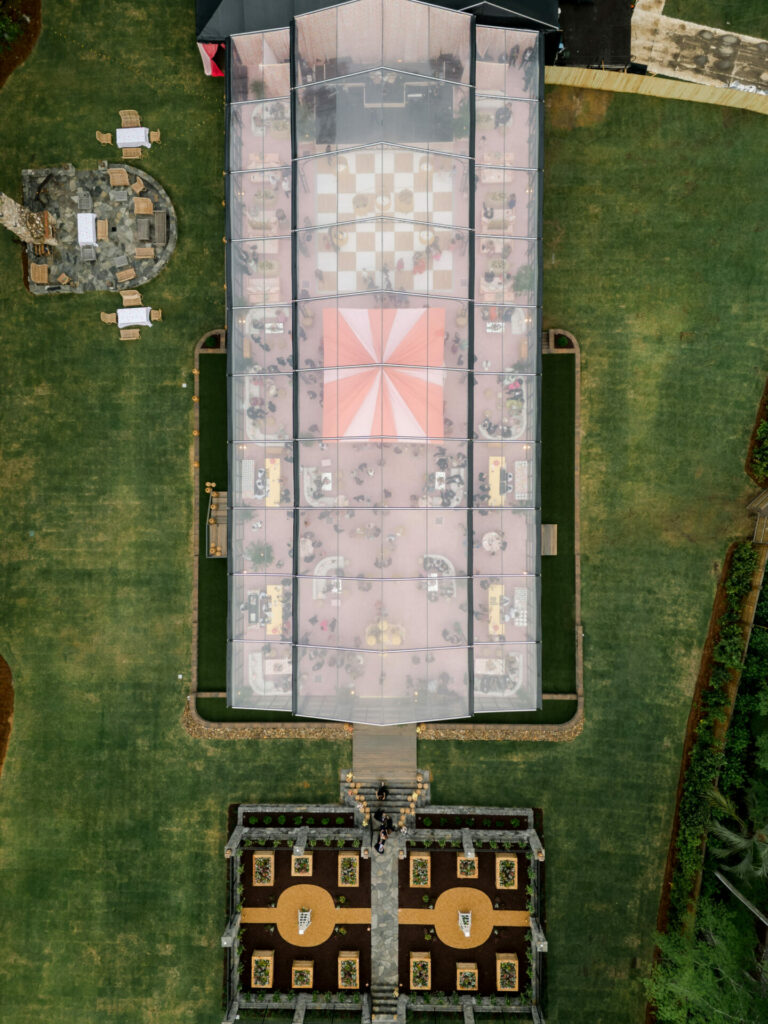Aerial view: transparent tent, tables, Katya Ruiz in gown, symmetrical garden.