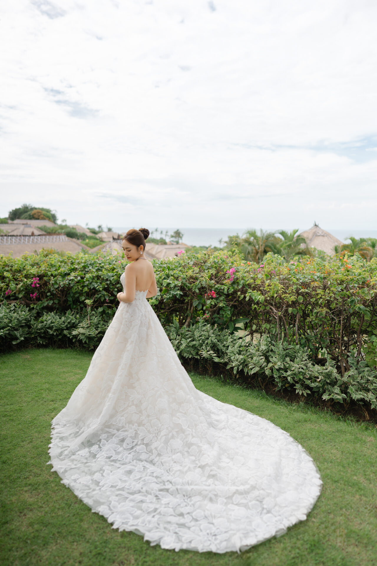 Bride in strapless gown stands on grass, scenic backdrop.