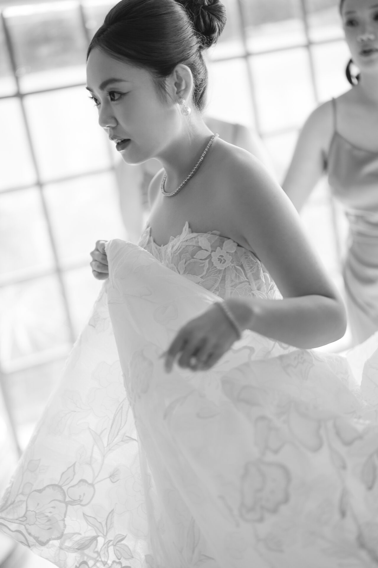 Bride holding dress, another woman behind, soft light.