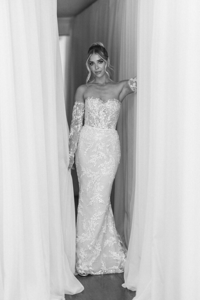 Bride in lace off-shoulder gown poses by sheer curtains.