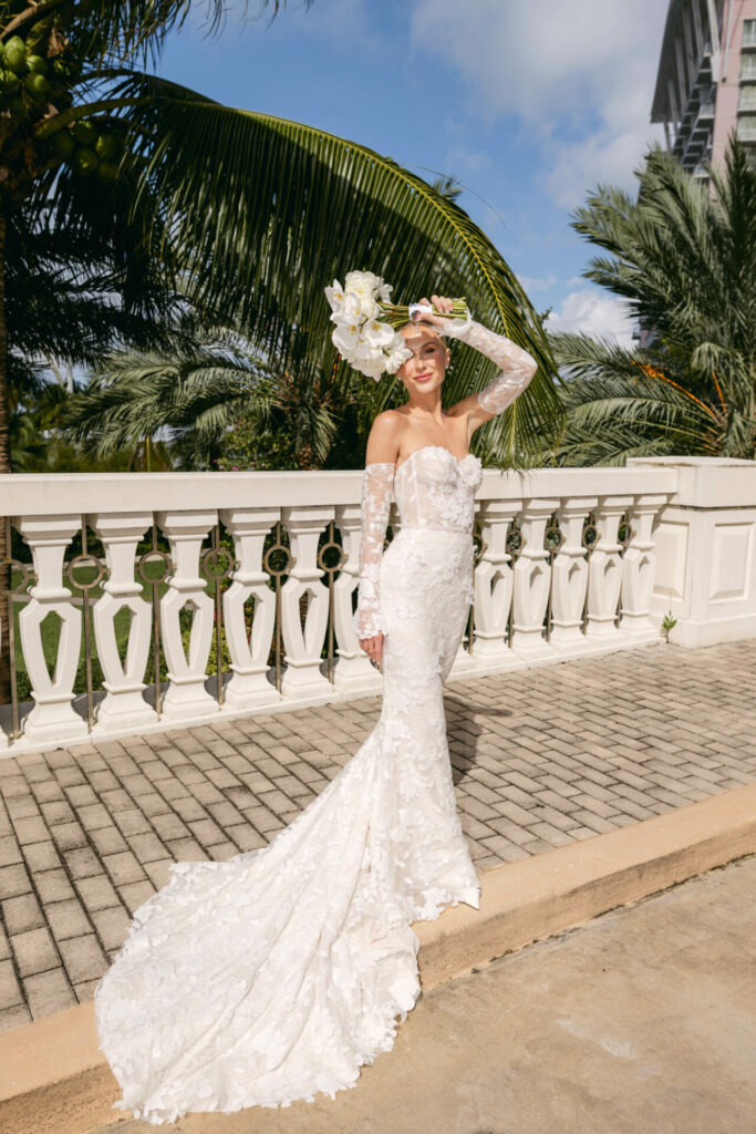 Bride in lace gown holds floral crown, smiling outside.