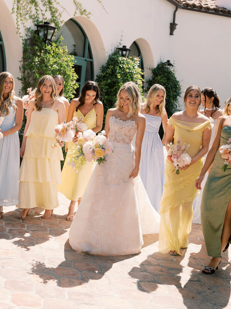 Bride and bridesmaids in pastel dresses smile outdoors.