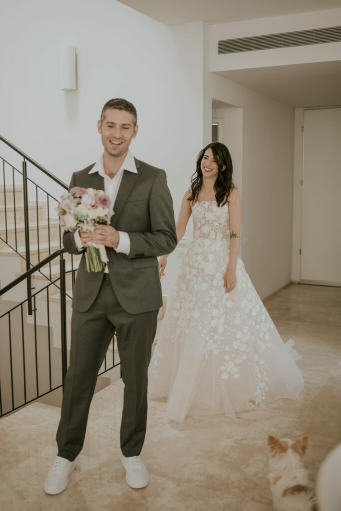 Man with flowers, bride approaches in couture gown; dog nearby.