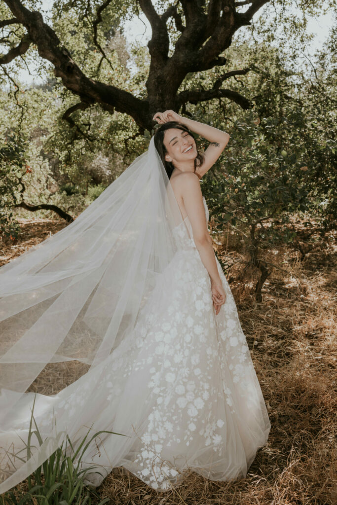 Katya Ruiz in a floral gown smiles outdoors among trees.