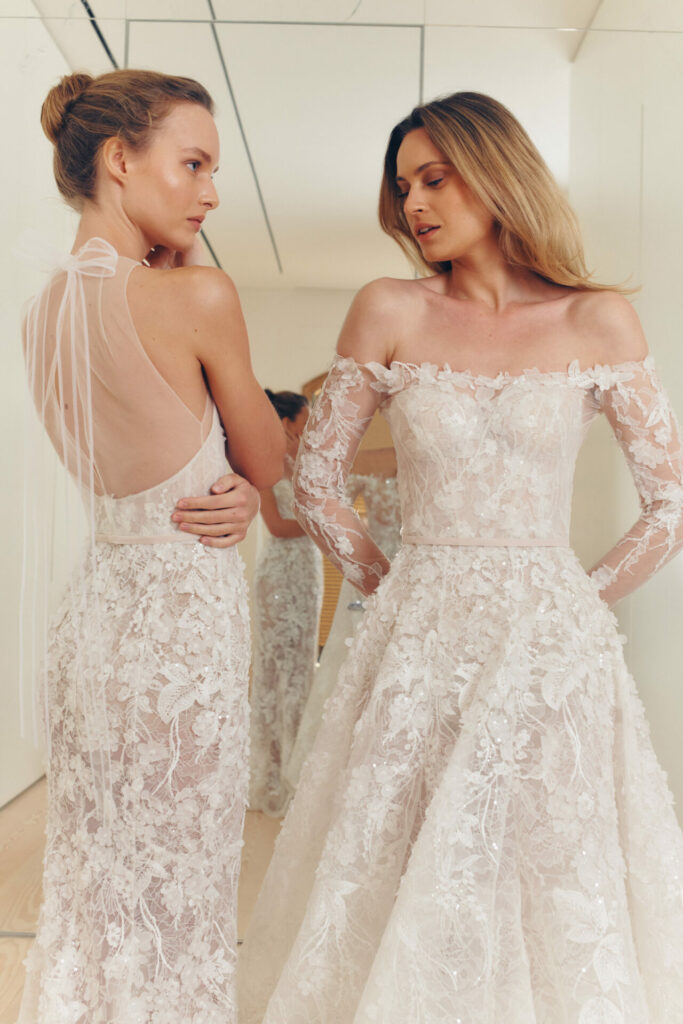Two women in white lace wedding dresses stand, gazing.