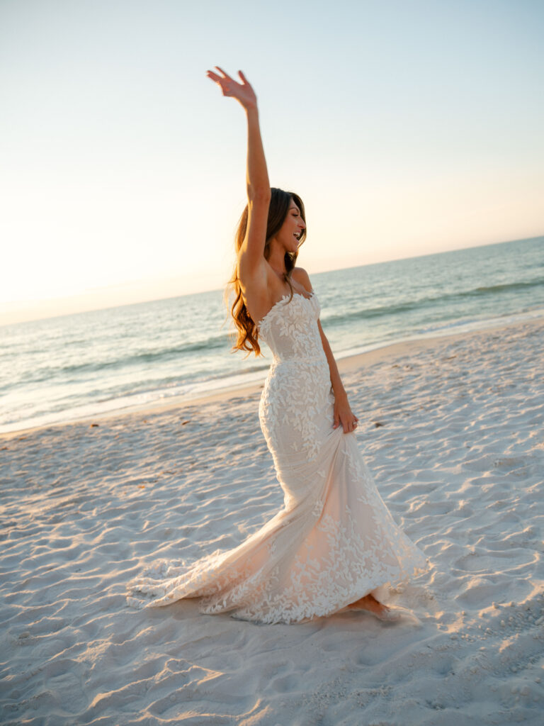 Bride in lace dress smiles, arm raised at sunset beach.