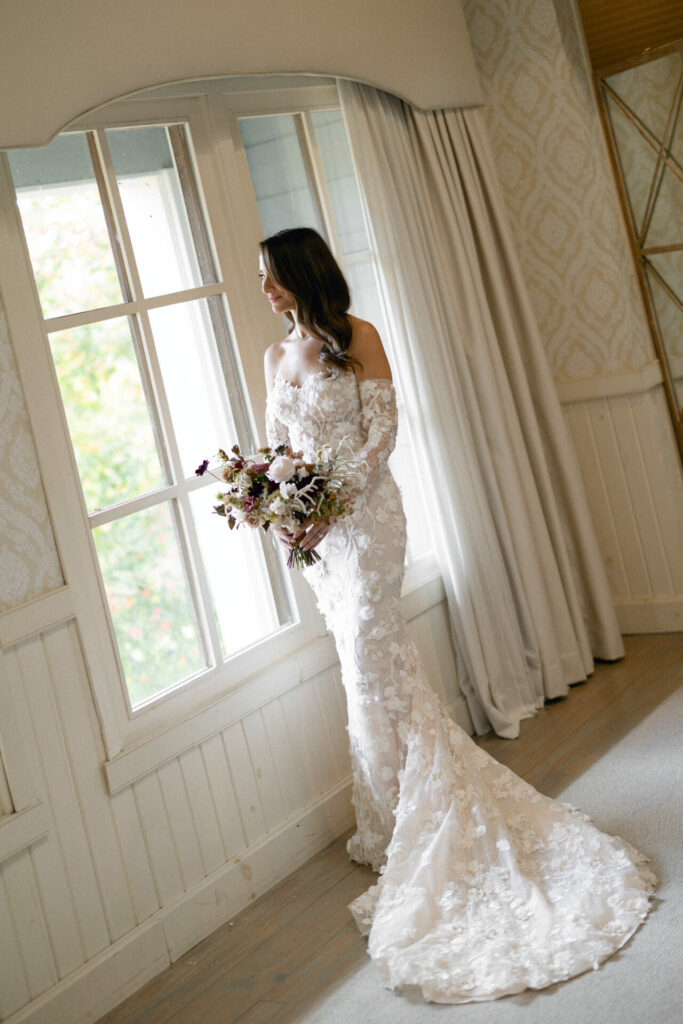Bride in SS2026 lace gown gazes out window, bouquet in hand.