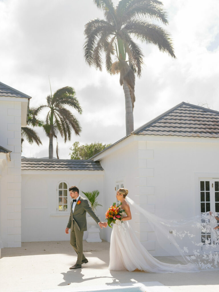 Bride and groom walk outside sunlit white building.