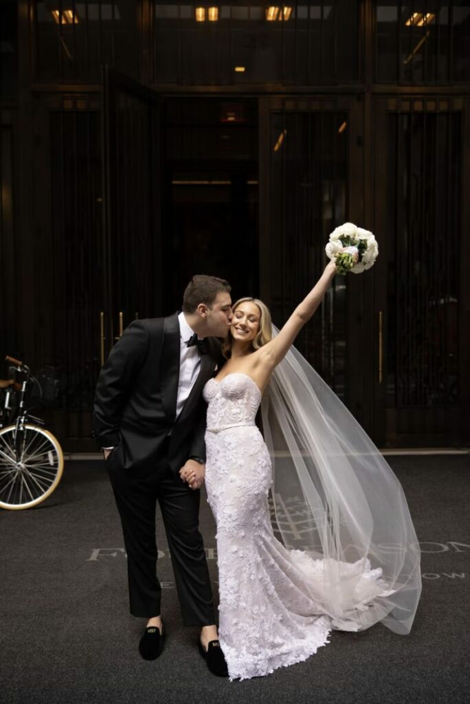 Bride in strapless gown kissed by groom, bouquet raised.