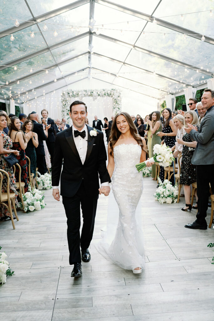 Bride and groom smile walking aisle as guests cheer.