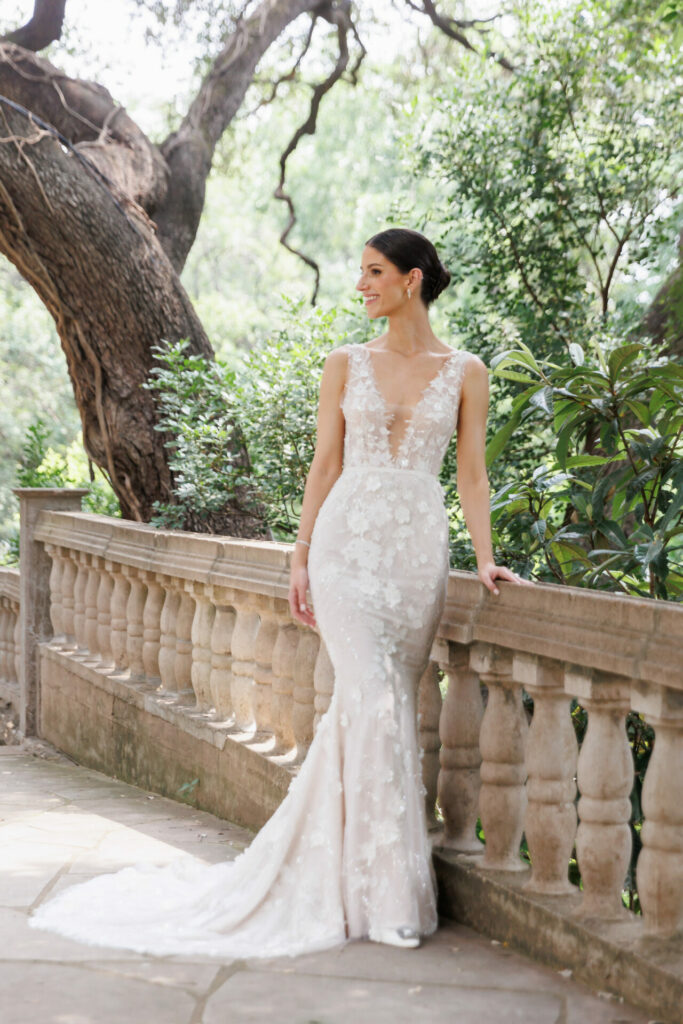 Bride in sleeveless lace gown smiles by stone railing outdoors.