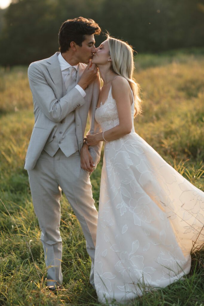 Bride and groom kiss in sunlit field, stylishly dressed.