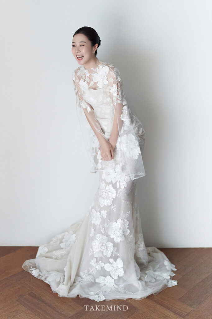 Woman in lace wedding gown poses, wooden floor visible.