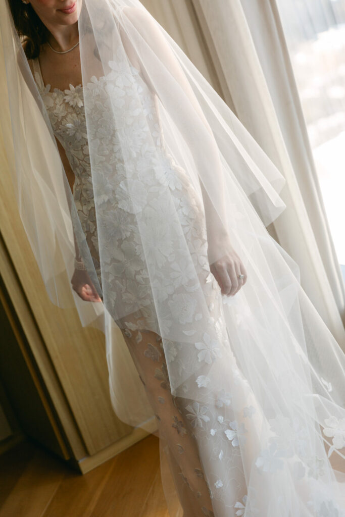 Bride in lace wedding dress by sunlit window, veiled.