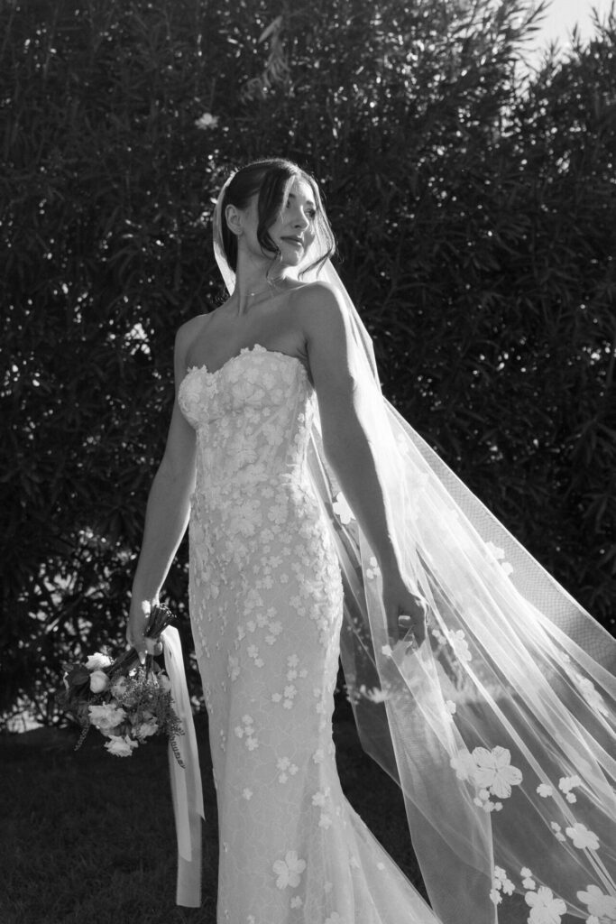 Bride in lace dress smiles outdoors, holding bouquet.