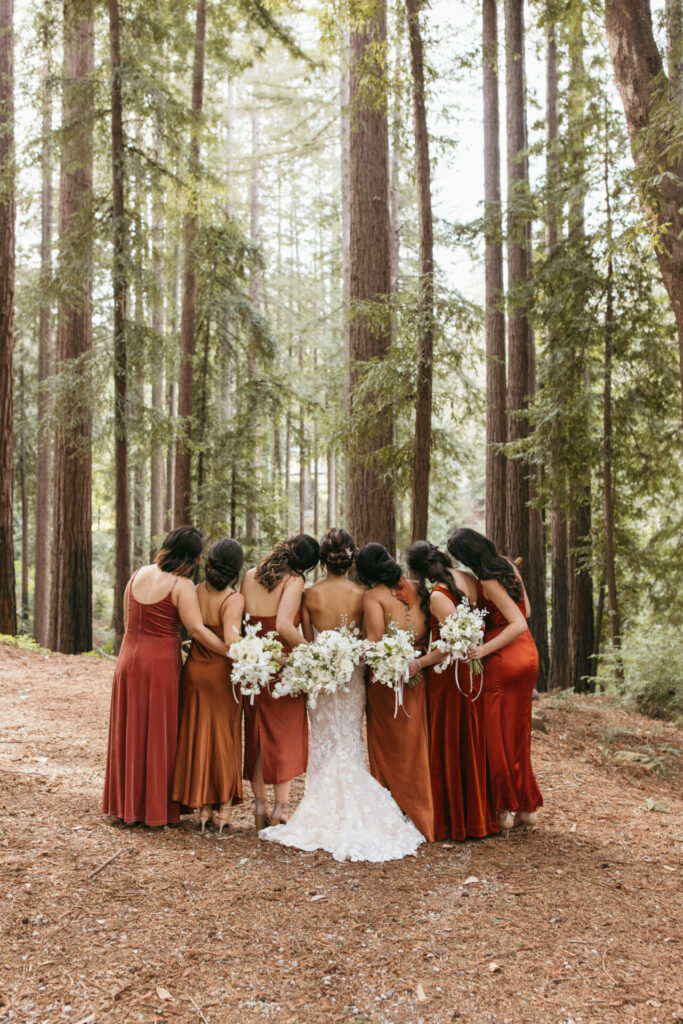 Bride and bridesmaids embrace with bouquets in redwood forest.