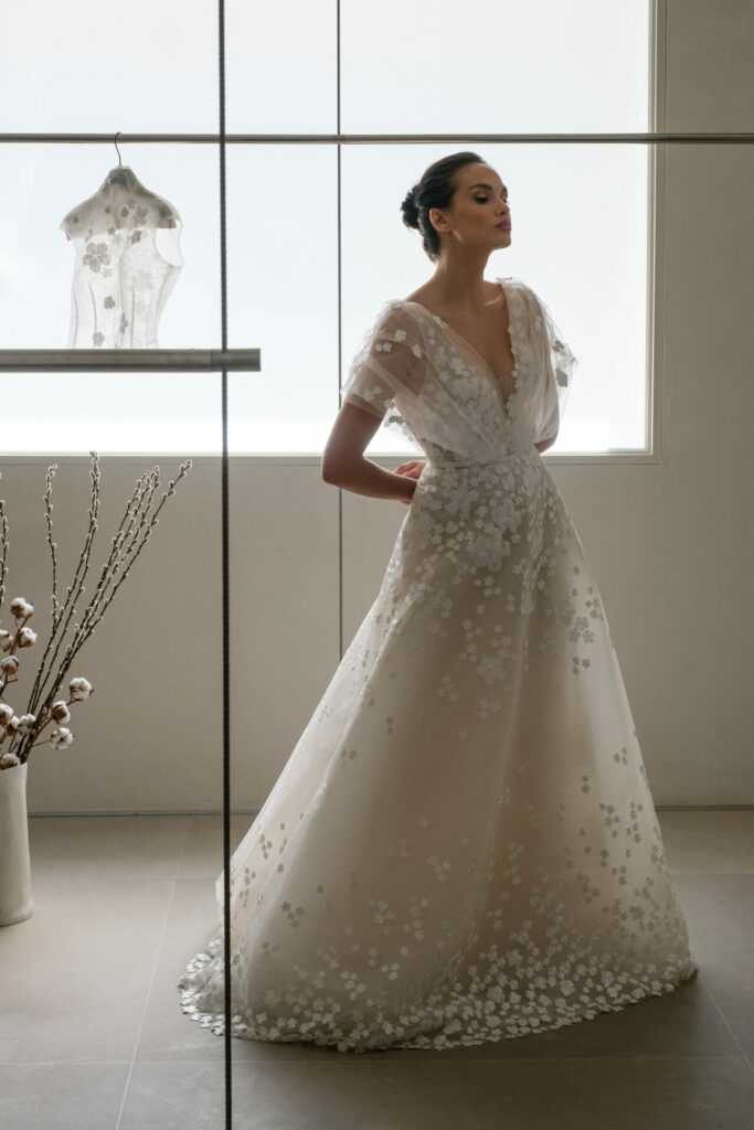 Woman in white sheer gown with bun, sunlight, top nearby.