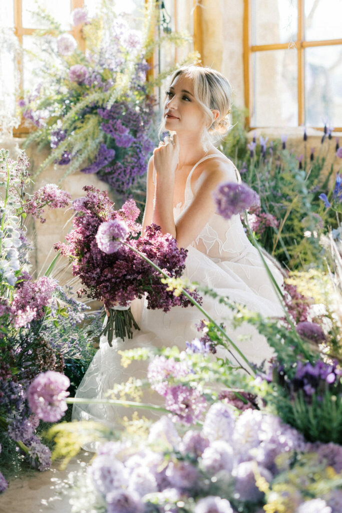 Woman in white dress amid purple flowers, soft sunlight.