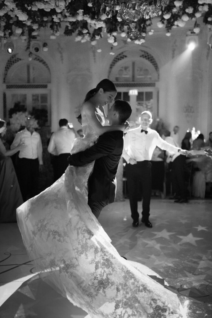 Groom lifts bride dancing, lace dress flows, guests watch.