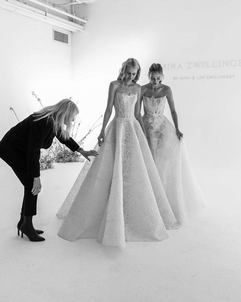 Two brides in elegant gowns; stylist adjusts a dress.