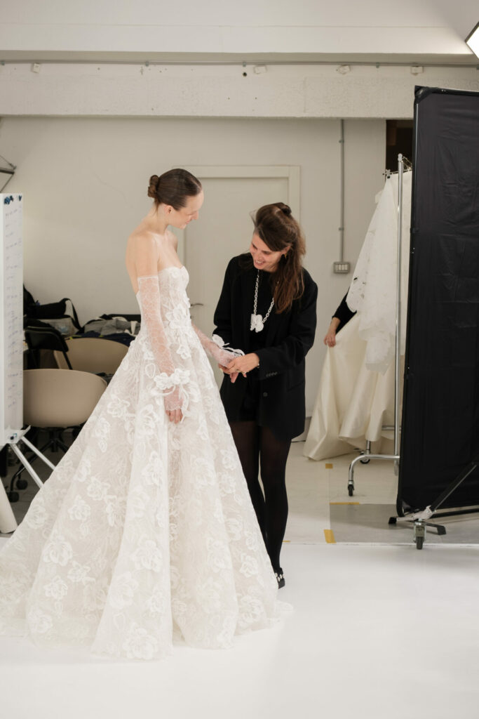 Bride in white lace gown, stylist adjusts dress.