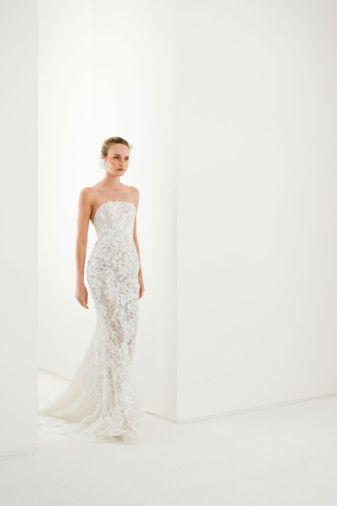 Woman in white lace gown stands calmly indoors.