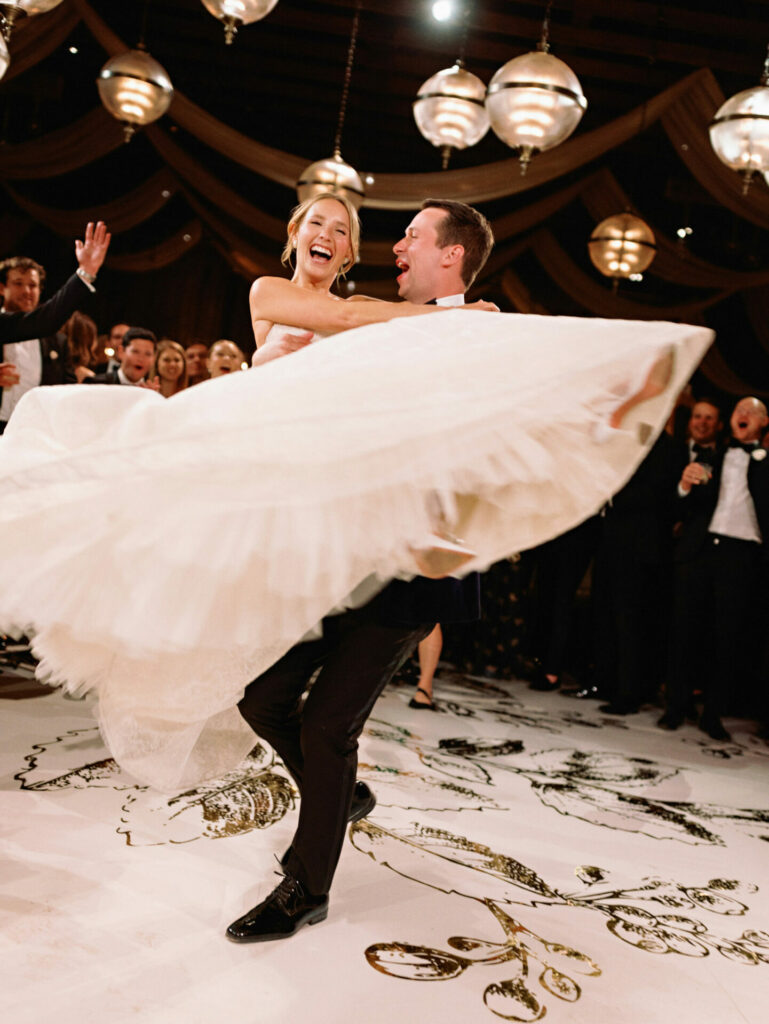 Groom twirls bride as her dress fans out, guests celebrating.