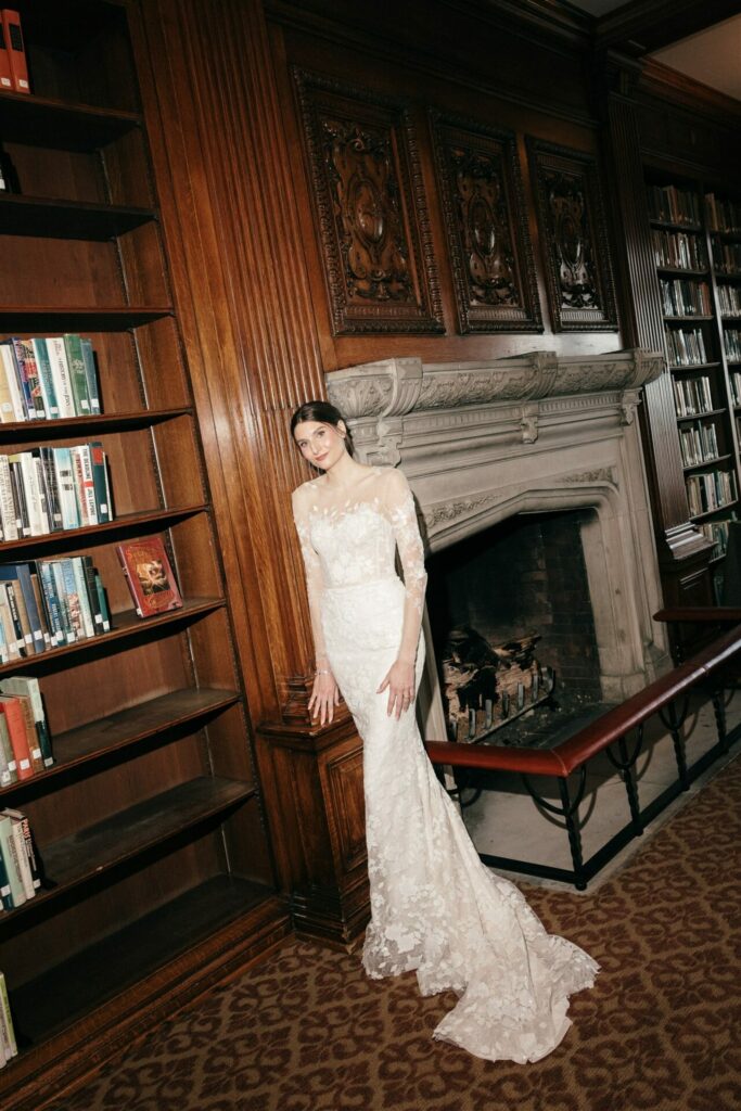 Bride in SS2026 lace gown stands by ornate library fireplace.