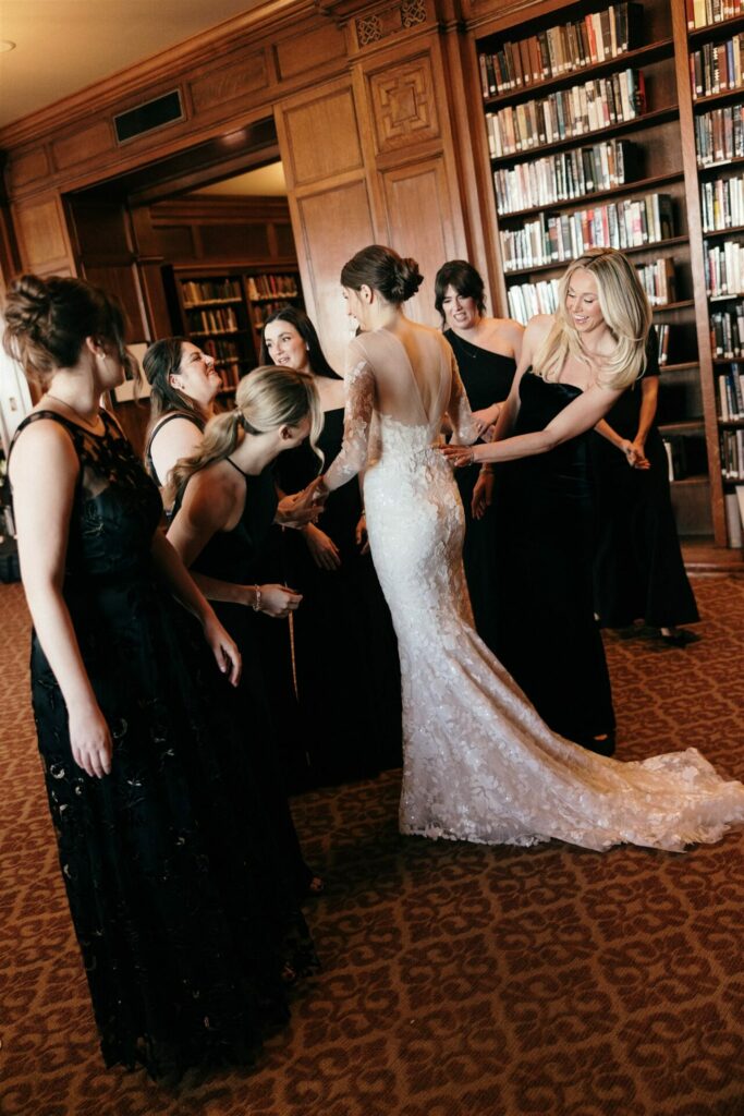 Bride in lace dress with bridesmaids in black gowns, library shelves behind.