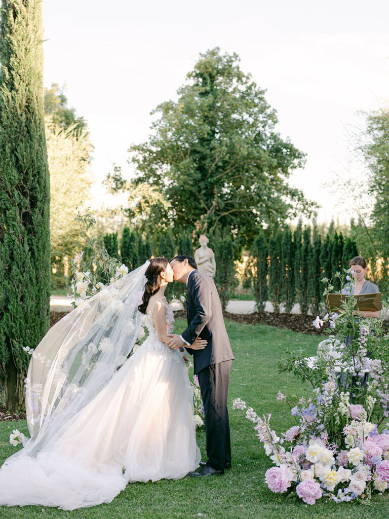 Bride and groom kiss outdoors amid flowers and greenery.