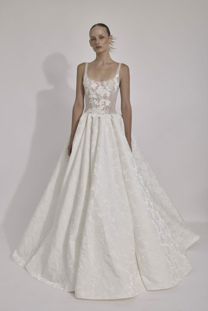 Woman in sleeveless white lace gown, plain background.