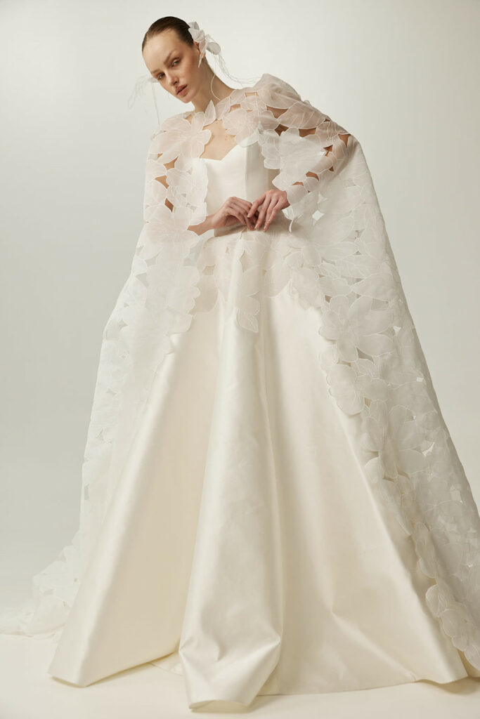 Woman in white satin gown, sheer cape, headpiece.